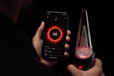 Image of a Puffco Peak Pro water piece that is black with red hue and hand holding and cellphone with a red ring and settings for the device.
