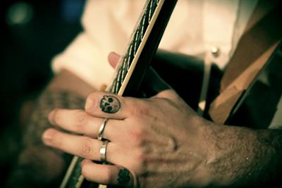 Hand on the neck of a guitar, with a tattoo of a skull on the pointer finger, and rings on the other fingers.