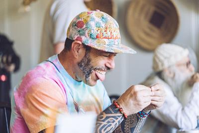 Man wearing with pastel tye dye t-shirt, arms tattooed, and floral baseball hat twisted up a joint in his hands with his tongue out.