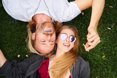 a woman with strawberry blonde hair and a man with blonde hair and facial hair share a joint while laying down in the grass