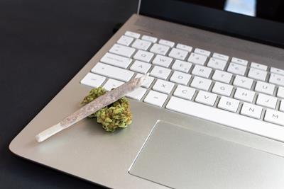 The corner of a laptop with a joint resting on top of a green nug on the space below the keyboard while someone is trying to be productive and high