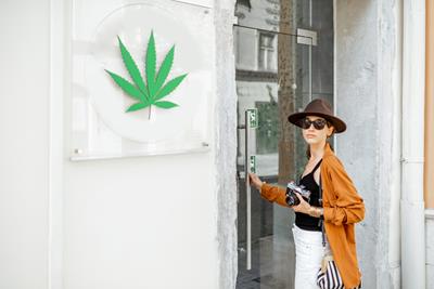 Tourist woman, with a camera around her neck, on an adventure for cannabis, about to open the door to a white dispensary with a weed sign out front 