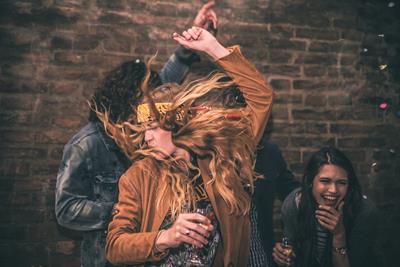 A group of friends that are dancing around in front of a brick wall. Women in front is whipping her blonde hair about with her hands in the air.