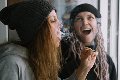 Two girls smoking a cannabis joint together with brown hair and blonde hair with beanies on top