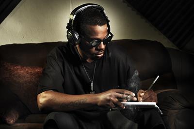 A black man is sitting down on a couch with black headphones on writing into a notebook