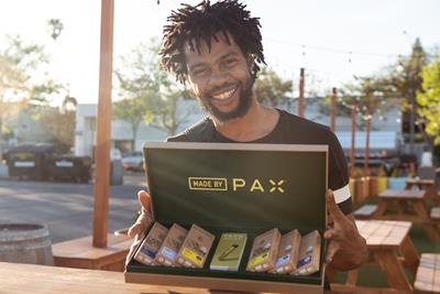 A black man with facial hair holding a box open ajar displaying  on the top of the lip "Made by Pax" and inside are an array of different cartridges packages that are in a beige boxes and have a green box in the center.